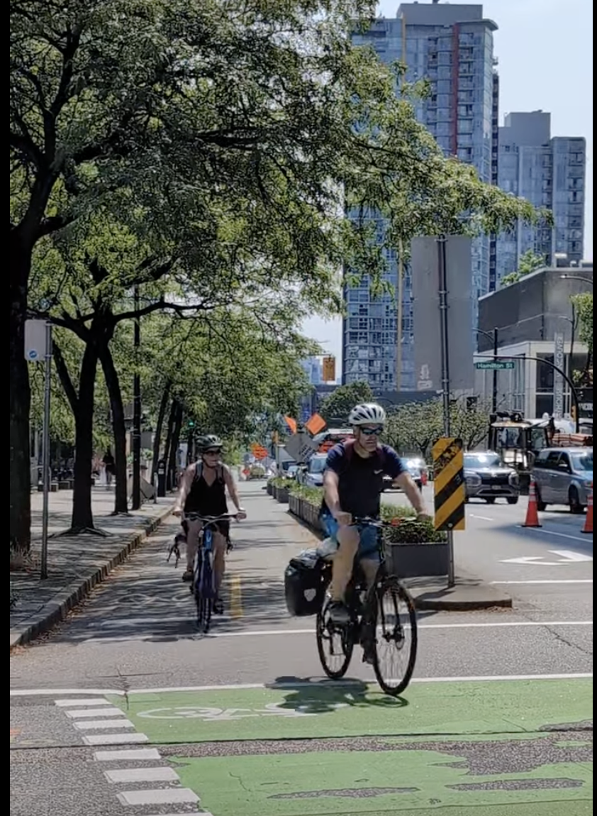 A protected bikeway with planted buffers and mature tree canopy on Smithe Street in Vancouver, British Columbia, with people biking and walking.